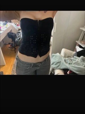 Svart korsett-topp med hålmönster - Säljer en svart korsett-topp med hålmönster och en liten slits framtill. Toppen är axelbandslös och har en tight passform som framhäver midjan. Perfekt för dig som vill ha en edgy och trendig look.