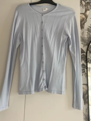 Ljusblå tunn kofta med knappar - Säljer en ljusblå tunn kofta med rund halsringning och långa ärmar. Koftan har knäppning framtill med små matchande knappar och är tillverkad i ett mjukt, tunt material som passar perfekt till lager på lager. Enkel och stilren design.
