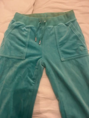 Turkosa velourbyxor Juicy Couture - Säljer ett par turkosa mjukisbyxor från Juicy Couture i mjuk velour. Dock haft brun utan sol i dom, vet ej om de går bort. Men de syns bara lite där längst upp som ni ser på bilden, mest inuti längst uppe.   Byxorna har bred mudd i midjan med dragsko, stora fickor fram och en ficka bak med knappar. Perfekta för en chill och trendig look.