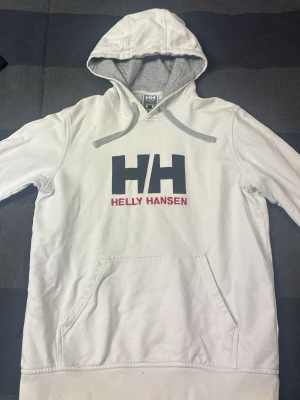 Vit hoodie från Helly Hansen - Säljer en vit hoodie från Helly Hansen med stor HH-logga och röd text på bröstet. Hoodien har grå dragsnören, mjuk insida och en stor magficka. M i storlek men passar S bättre