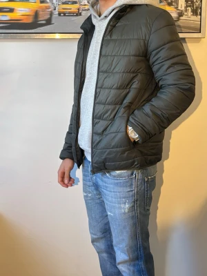 Grön pufferjacka med dragkedja - Säljer en grön pufferjacka med dragkedja och sidofickor. Jackan har en stilren, quiltad design och är lätt vadderad för extra värme. Perfekt för kalla dagar och enkel att matcha med olika outfits. Passar dig som gillar en clean och modern look.