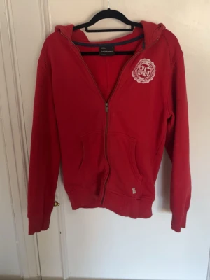 Röd zip hoodie från Peak Performance - Snygg röd hoodie med dragkedja från Peak Performance. Har huva, två fickor framtill och ett vitt tryckt logomärke på bröstet. Tillverkad i mjukt bomullsmaterial som känns skönt mot huden. Perfekt för en avslappnad och sportig stil.