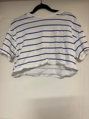 Vit och blå randig croppad topp - Snygg croppad topp i vitt med blåa ränder. T-shirten har korta ärmar och en avslappnad passform, perfekt till jeans eller kjol. Tillverkad i mjuk bomull för en skön känsla.