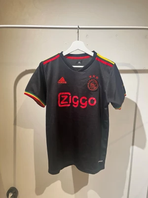 Ajax svart matchtröja Adidas - Säljer en svart Ajax matchtröja från Adidas med Ziggo-logga på bröstet och klubbmärke med tre stjärnor. Tröjan har färgglada detaljer i rött, gult och grönt på axlar och ärmslut samt texten 'CURA' på ena ärmen. Tillverkad i funktionsmaterial, perfekt för fotboll. SÄLJS INTE LÄNGRE.  