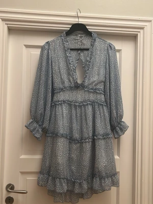 Blå volangklänning med mönster - Supersöt blå klänning med småvitt blommönster, volanger och v-ringning. Klänningen har långa puffärmar med resår vid ärmslut och en öppen detalj i ryggen. Materialet är tunt och lätt, troligen polyester eller liknande. Perfekt för dig som gillar romantisk och trendig stil.