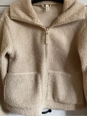 Beige teddyjacka från H&M - Mysig beige teddyjacka från H&M med dragkedja framtill och två stora fickor. Jackan har hög krage och är gjord i ett fluffigt, mjukt material som håller dig varm. Perfekt för kyliga dagar och enkel att matcha med olika outfits.