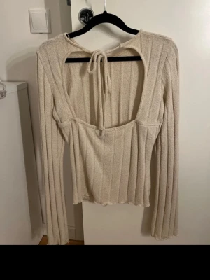 Beige ribbad topp med öppen rygg - Trendig beige ribbad topp med lång ärm och snygg öppen rygg med knytband i nacken. Toppen har en croppad passform och är tillverkad i ett mjukt, stretchigt material som känns skönt mot huden. Perfekt för dig som gillar stilrena detaljer och en modern look.