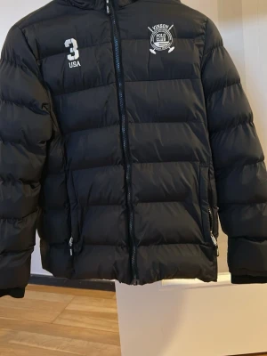 Svart dunjacka Vinson Polo Club - Snygg svart dunjacka från Vinson Polo Club med huva och dragkedja framtill. Pris diskuteras Storleken är 175-178 cm