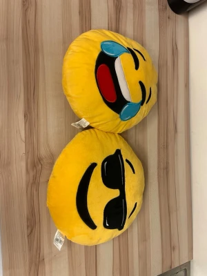 Emoji-kuddar i gult tyg - Två runda emoji-kuddar i mjukt gult tyg. Ena kudden har ett skrattande ansikte med tårar och den andra har ett ansikte med svarta solglasögon. Perfekta som dekoration i soffan eller på sängen. Helt nya!