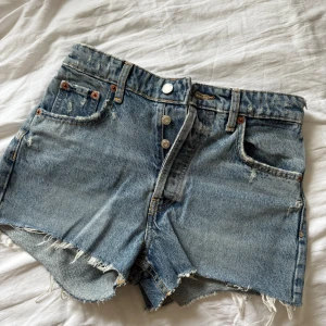 Blå jeansshorts med rå kant - Klassiska blå jeansshorts med mid midja och rå, fransig kant. Shortsen har fem fickor, bälteshällor och stängs med knapp. Perfekta för varma dagar och en avslappnad stil.