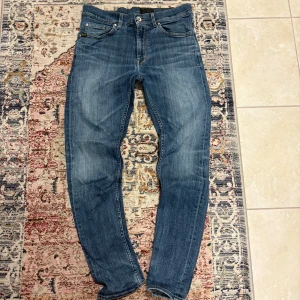 Blå slim fit jeans från Tiger of Sweden - Säljer ett par klassiska blå slim fit jeans från Tiger of Sweden med fem fickor och snygga slitningar. Jeansen har normal midja, dragkedjegylf och bruna sömmar. Jag säljer då de inte passar längre. Finns inga defekter.
