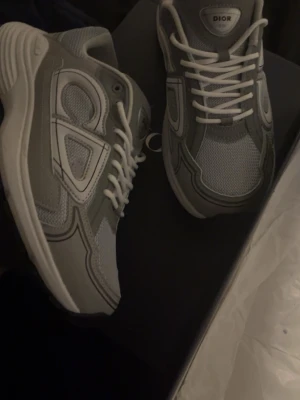 Grå Dior sneakers med vita detaljer - Snygga grå sneakers från Dior med vita detaljer och vita skosnören. Skorna har en modern design med flera lager mesh och syntetmaterial, samt en tjock vit sula för extra komfort. Perfekta för dig som gillar exklusiva och trendiga sneakers. Fick som present har inte kommit till någon användning 