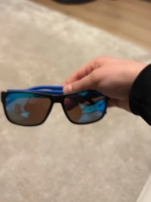 Svarta solglasögon med blå detaljer - Snygga solglasögon med svart båge och blå detaljer på skalmarna. Glasen har en spegelblå ton som ger en cool look. Perfekta för att lyfta din stil under soliga dagar.Tommy Hilfiger 