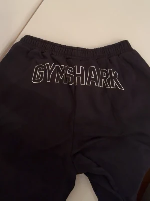 Svarta mjukisbyxor från Gymshark - Säljer ett par svarta mjukisbyxor från Gymshark med elastisk midja och tryckt logga bak samt texten 'STRENGTH DEPT.' fram. Sköna och avslappnade byxor i mjukt material, perfekta för chill eller träning.
