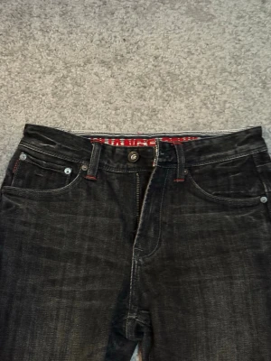 Svarta raka jeans - Säljer ett par svarta jeans med raka ben och snygga röda sömmar vid fickorna. Jeansen har klassisk femficksmodell och knappgylf. Materialet är denim och insidan av linningen har ett rött mönster. Perfekta för en cool och avslappnad stil. Osäker på märket. 
