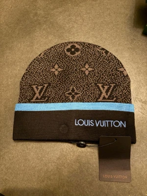 Loui ton mössa - Snygg mössa från Loui ton i brunt med klassiskt monogrammönster och blå kant. Märkets logga är broderad i ljusblått framtill. Tillverkad i mjukt stickat material som känns lyxigt och trendigt. Perfekt accessoar för dig som vill sticka ut.