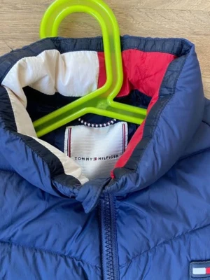 Blå dunväst från Tommy Hilfiger - Snygg blå dunväst från Tommy Hilfiger med quiltad design och dragkedja framtill. Västen har ståkrage och innerfoder i vitt och rött, samt det klassiska Hilfiger-logotypen på bröstet. Perfekt för lager på lager och streetwear-stil. Fast pris!