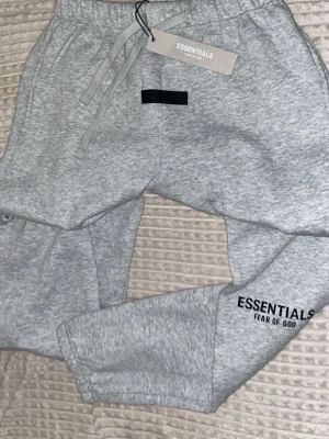 Grå Essentials Fear of God mjukisbyxor storlek M - Snygga grå mjukisbyxor från Essentials Fear of God med logga tryckt på benet. Byxorna har dragsko i midjan och muddar vid bensluten för en skön och avslappnad passform. Perfekta för chill dagar och streetwear-stil.