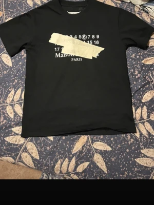 Svart Maison Margiela t-shirt - Svart t-shirt från Maison Margiela med ikoniskt tryck framtill i vitt och beige, där siffror och text delvis täcks av tejpdesign. Klassisk passform och rund halsringning. Perfekt för dig som gillar streetwear och unika designer.