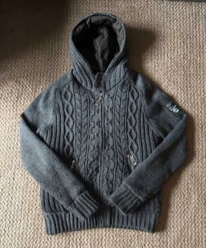 Stickad cardigan - Säljer en mörkgrå stickad hoodie  med snyggt kabelstickat mönster framtill och ribbad baksida. Tröjan har huva med quiltad insida, två fickor med dragkedja och långa ärmar. Perfekt för dig som gillar chill och stil på samma gång.