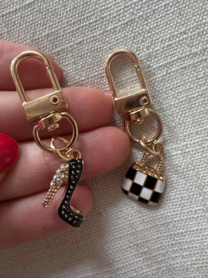 Två nyckelringar - Två guldiga nyckelringar med hängande charms: en svart stilettklack dekorerad med små stenar och en svartvit rutmönstrad väska. Har bundlerabatt vid köp av fler varor🥰