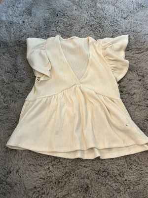 Krämvit blus med volangärm - Supersöt krämvit blus med djup v-ringning och volangiga korta ärmar. Blusen har en lätt rynkad midja och ett mjukt, ribbat tyg som ger en avslappnad men ändå chic look. Perfekt att styla med jeans eller kjol för en trendig outfit.
