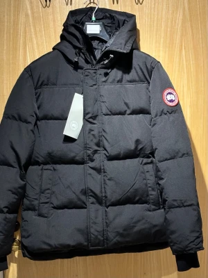 MacMillan jacka från Canada Goose - (L i eu storlek) Säljer en svart pufferjacka från Canada Goose med huva och klassisk logotyp på ärmen. Jackan har dragkedja och knäppning framtill, två sidofickor och ribbade muddar vid ärmsluten. Perfekt för kalla vinterdagar och riktigt snygg streetstil.