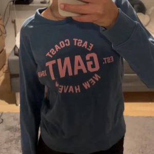 Blå sweatshirt från GANT - Säljer en blå sweatshirt från GANT med tryck i rosa på bröstet där det står 'EAST COAST GANT NEW HAVEN'. Tröjan har rund halsringning och ribbade muddar vid ärmslut och nederkant. Perfekt för en avslappnad och sportig stil.