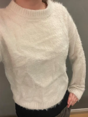 Vit fluffig stickad tröja - Mysig vit stickad tröja med fluffig yta. Tröjan har långa ärmar och ribbade muddar vid ärmslut och nederkant. Perfekt för kalla dagar när du vill vara både varm och stilren. Lite sliten efter tvätt men fortfarande skön. Ej använt på väldigt länge. Ej bra pris,, skriv till mig