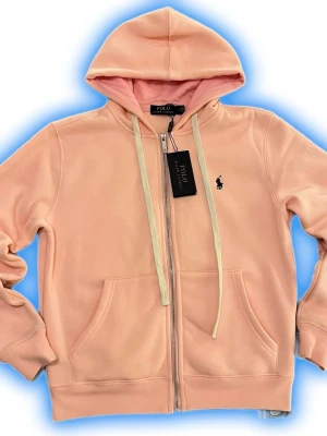 Ljusrosa hoodie från Polo Ralph Lauren - Snygg ljusrosa hoodie från Polo Ralph Lauren med dragkedja framtill, vita snören och klassisk huva. Broderad logga på bröstet och två stora fickor fram. Tillverkad i mjuk bomullsblandning som är jätte skön mot huden. Perfekt för en chill och avslappnad stil.