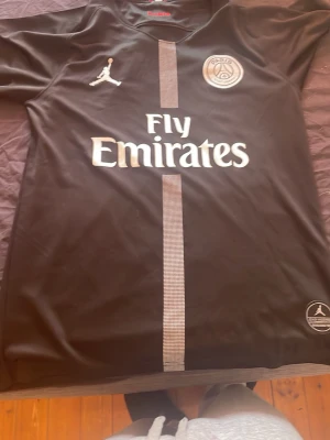 Svart PSG x Jordan matchtröja S - Snygg svart PSG matchtröja i samarbete med Jordan. Tröjan har vita detaljer, PSG-logga och Fly Emirates-tryck på bröstet samt en vertikal rand i mitten. Tillverkad i lätt och ventilerande material, perfekt för fotboll eller träning. Pris kan diskuteras 