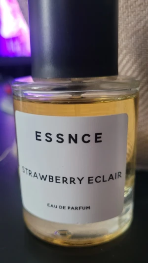 ESSNCE Strawberry Eclair parfym - En stilren parfymflaska från ESSNCE med doften Strawberry Eclair. Flaskan är genomskinlig med en svart cylinderformad kork och vit etikett med svart text. Innehållet är ljusgult och parfymen är en Eau de Parfum.