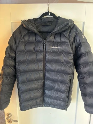 Svart dunjacka Peak Performance M - Snygg svart dunjacka från Peak Performance med huva och dragkedja framtill. Jackan har två sidofickor med dragkedja och diskret logga på bröstet. Perfekt för kalla vinterdagar och riktigt skön att bära. Klassisk puffer-modell som alltid är trendig.