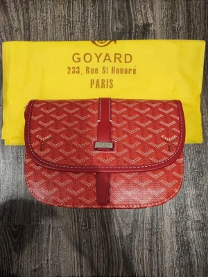 Röd axelremsväska  - Snygg röd axelremsväska med klassiskt mönster i vitt och beige. Väskan har en elegant rektangulär form, lock med magnetstängning och detaljer i rött läder. Perfekt för dig som vill sticka ut med en ikonisk accessoar.