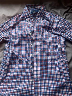 Rutig skjorta från Ralph Lauren - Klassisk rutig skjorta från Ralph Lauren i blått, rött och vitt. Skjortan har knappar framtill, button-down krage och den ikoniska broderade loggan på bröstet. Tillverkad i mjuk bomull och perfekt för en avslappnad men snygg stil.