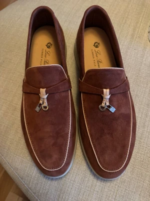 Bruna mocka loafers från Loro Piana - Snygga bruna loafers i mocka från Loro Piana med vita kontrastsömmar och dekorativ metall-detalj framtill. Skorna har en klassisk form och ljus innersula med logotyp. Perfekta för en stilren och avslappnad look. Inget kvitto därav priset.  