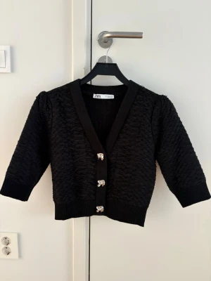 Svart kort kofta från Zara - Säljer en svart kort kofta från Zara med strukturstickat tyg och tre dekorativa knappar framtill. Koftan har 3/4-ärmar, bred ribbad kant och v-ringad hals. Perfekt för att lyfta en enkel outfit.