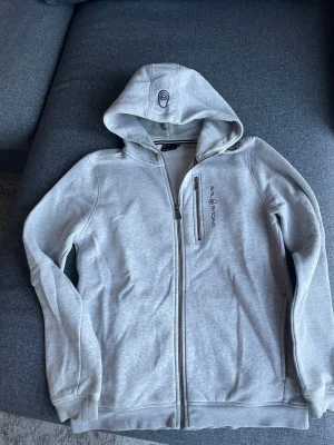 Ljusgrå ziphoodie från Sail Racing stl170  - Ljusgrå hoodie från Sail Racing med hel dragkedja och huva. 