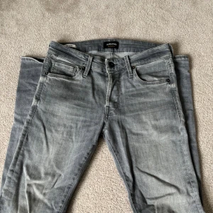 Grå slim fit jeans från Jack & Jones - Säljer ett par grå slim fit jeans från Jack & Jones med klassisk femficksdesign och svarta nitar. Jeansen har en snygg tvättad look och normal midja. Perfekta för dig som gillar en modern och avslappnad stil.