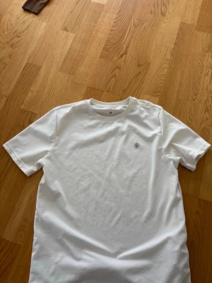 Vit t-shirt från Lumberjack - Klassisk vit t-shirt från Lumberjack med rund hals och diskret logga på bröstet. Tillverkad i mjuk bomull för en skön känsla och enkel stil. Perfekt till jeans eller shorts för en clean look.