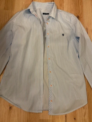 Blårandig skjorta från Marc O'Polo - Snygg blårandig skjorta från Marc O'Polo med klassisk krage och vita knappar med röda detaljer. Skjortan har lång ärm och diskret logga broderad på bröstet. Tillverkad i mjuk bomull för en bekväm känsla.