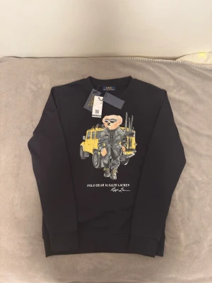 Svart Polo Bear hoodie Ralph Lauren - Svart hoodie från Ralph Lauren med Polo Bear-tryck framtill. Björnen har solglasögon och står framför en gul bil. Klassisk passform med ribbade muddar och rund hals. Perfekt för dig som gillar streetstyle och coola prints.