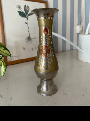 Metallvas med blommönster - Säljer en dekorativ metallvas i silver och mässingston med handmålat blommönster i rött och vitt. Vasen har en smal midja och vid öppning, perfekt som inredningsdetalj eller för enstaka blommor. Unik vintagekänsla och snygg form.