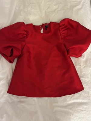Röd puffärm topp från Vero Moda - Trendig röd topp från Vero Moda med stora puffärmar och rund halsringning. Toppen har en söt knytning i nacken och är tillverkad i ett glansigt material som ger en lyxig känsla. Perfekt för dig som vill sticka ut med en färgstark look. Aldrig använd 