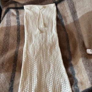 Lång stickad kjol med snörning - Säljer en lång, ljus stickad kjol i offwhite med dekorativt snörningsband i midjan och virkade detaljer. Kjolen har en tajt passform och är perfekt att ha över en bikini