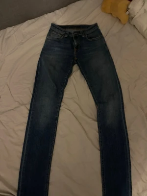 Blå smala jeans från Nudie Jeans - Säljer ett par klassiska blå jeans från Nudie Jeans med smal passform och snygga slitningar. Jeansen har fem fickor, orange sömmar på bakfickorna och är tillverkade i denim. Perfekta för dig som gillar en tidlös och avslappnad stil. Hör av er vid frågor. Pris går att diskutera