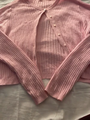 Rosa ribbad kofta från 157 - Söt ljusrosa ribbad kofta från 157 med rund halsringning och knappar framtill. Koftan har långa ärmar och är tillverkad i ett mjukt, stretchigt material som sitter skönt på kroppen. Perfekt att slänga över en topp för en chill look.