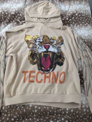 Beige hoodie med tiger och TECHN0-print - Säljer en beige hoodie med stor tigertryck och texten TECHN0 framtill. Ärmarna har detaljer i guldmönster som liknar fjädrar eller vingar. Huvan har dragsko och tröjan är i mjukt bomullsliknande material. Perfekt för dig som gillar statement-plagg och unika prints.
