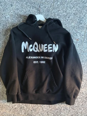 Svart hoodie från Alexander McQueen - Snygg svart hoodie från Alexander McQueen med stor vit logga och texten 'EST. 1992' på bröstet. Klassisk känguruficka framtill, huva med dragsko och ribbade muddar. Tillverkad i Italien, mjukt och bekvämt material som passar perfekt till streetwear-stilen. Mer info eller bilder Skirv 