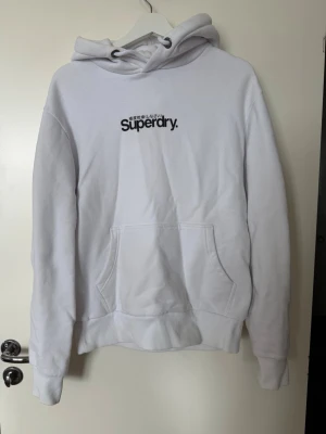 Vit hoodie från Superdry - Säljer en clean vit hoodie från Superdry med svart logga och japansk text på bröstet. Hoodien har stor känguruficka, ribbade muddar och en rejäl huva med dragsko. Perfekt oversized vibe och mjukt material, troligen bomullsmix.
